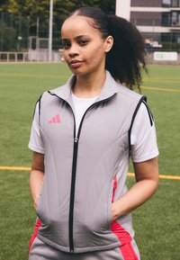 Pilkas sportinis liemenė su rožiniais akcentais ir užtrauktuko uždarymu, derinamas su balta marškinėliais. Lygi medžiaga, skirta sportui, turi šoninius kišenes.