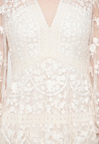 Gros plan sur une robe en dentelle blanche avec motifs floraux, encolure en V et longues manches transparentes ornées de fleurs brodées.