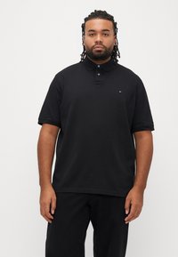 Čierna polokošeľa s klasickým golierom, krátkymi rukávmi a gombíkovým zapínaním. Na hrudi má diskrétnu značku. Mäkká, hladká textúra.