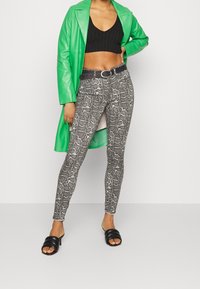 Top corto de canalé negro, abrigo largo de cuero verde, jeans ajustados de serpiente en tonos grises, cinturón negro y sandalias negras de dedo.