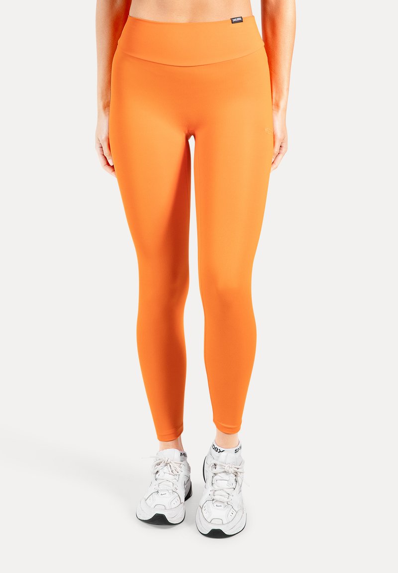 Collants orange vif avec une taille haute, confectionnés dans un matériau extensible. Portés avec des chaussures de sport blanches. Texture lisse, coupe ajustée.