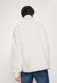 Giacca in pile bianca con colletto alto, vestibilità oversize e orlo leggermente arrotondato. Presenta una texture morbida e polsini elasticizzati.