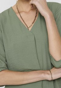 Blusa in verde oliva, testurizzata, con scollatura a V. Presenta maniche corte, una leggera caduta e un design a strati. Collana semplice in oro sottolinea l'outfit.
