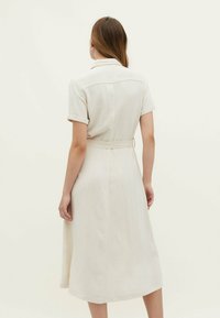 Robe midi en lin beige avec des manches courtes roulées, un col et une ceinture à la taille. Présente un léger motif texturé et un ourlet évasé.