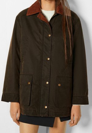 Femme portant une veste marron foncé avec col en velours côtelé, boutons-pression et grandes poches avant, par-dessus un pull blanc et une jupe noire.