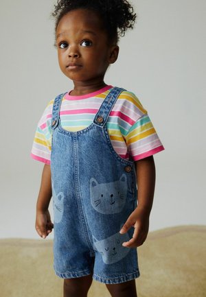 Småbarn iført denim-overalls med katteansigter og en t-shirt med stribet regnbuemønster, stående foran en ensfarvet lys baggrund.