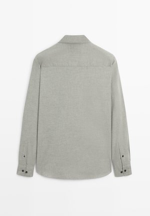Chemise à manches longues gris clair avec une texture lisse, un col pointu et des poignets boutonnés. Présente un empiècement au dos et un ourlet arrondi.