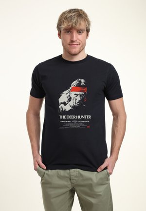 Sort bomulds T-shirt med grafisk tryk af en mand med et rødt pandebånd, tekst med ordene "THE DEER HUNTER" i hvid og grå.