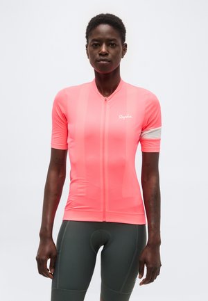 Mujer con un maillot de ciclismo ajustado de color rosa brillante con mangas cortas y pantalones cortos de ciclismo acolchados de color gris oscuro, de pie sobre un fondo liso.