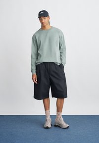 Felpa verde texture, pantaloni corti neri e sneakers grigie con accenti. Il modello indossa un cappellino nero e calze chiare. Design semplice e vestibilità casual.