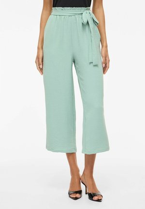 Kobieta ubrana w miętowozielone, wysokiej talii, skrócone spodnie o szerokich nogawkach z wiązanym paskiem z tkaniny oraz czarne sandały na otwartym palcu na białym tle.