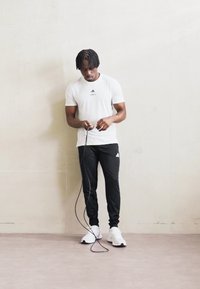 Bílá tričko s krátkým rukávem Adidas, černé sportovní kalhoty a bílé tenisky. Osoba drží švihadlo na neutrálním pozadí.