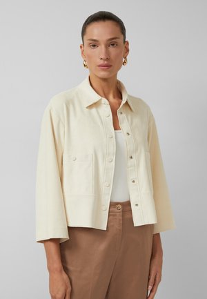 Vrouw draagt een beige jasje met knopen over een wit topje, met een bruine broek met hoge taille en gouden oorbellen in hoepelvorm tegen een effen achtergrond.