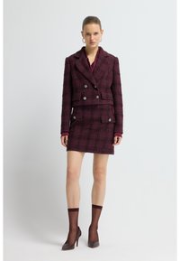Costume en tweed bordeaux avec une veste croisée, des finitions contrastantes et un motif à carreaux. Assorti d'une jupe coordonnée et de talons bruns.