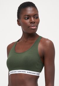 Soutien-gorge de sport vert olive avec une texture lisse, une coupe ajustée et une bande élastique blanche arborant le logo "BJÖRN BORG" en bas.