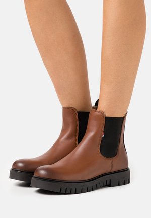 Bottines en cuir marron avec des panneaux latéraux élastiques noirs et de épaisses semelles crantées noires, portées sur des jambes nues.