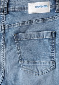 Jean en denim bleu clair avec une poche arrière et une étiquette blanche portant l'inscription "PAS DE SOUCIS JUSTE DU BONHEUR." Les coutures sont visibles dans une couleur contrastante.