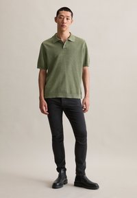 Młody mężczyzna stojący w zielonej koszulce polo z krótkim rękawem, czarnych dopasowanych dżinsach i czarnych botkach za kostkę na jednolitym beżowym tle.