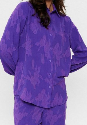 Femme portant une chemise violette à manches longues boutonnée et un pantalon assorti avec un motif floral subtil.