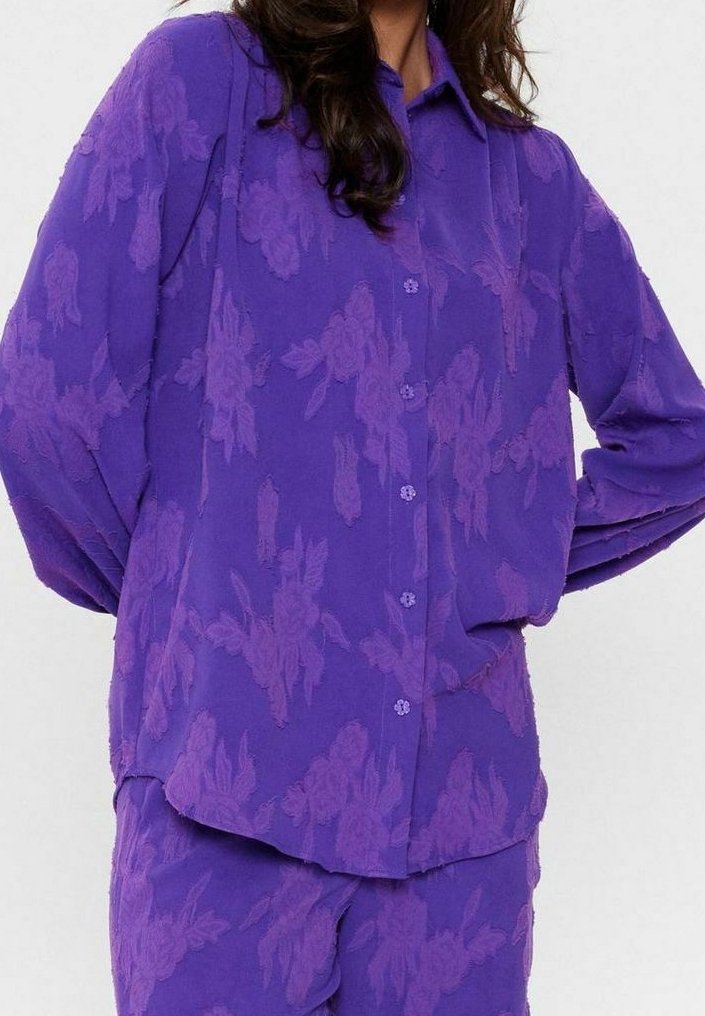 Femme portant une chemise violette à manches longues boutonnée et un pantalon assorti avec un motif floral subtil.