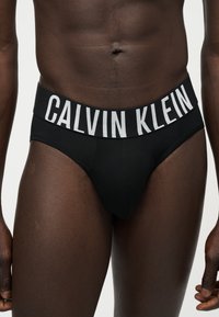 Άνδρας με τούρσο που φοράει μαύρα εσώρουχα με φαρδύ λάστιχο μέσης με μεγάλα λευκά γράμματα "Calvin Klein" σε μονόχρωμο φόντο.
