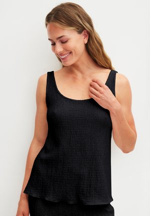 Vrouw met lang haar die een gestructureerde zwarte mouwloze top draagt, glimlacht en naar beneden kijkt, hand die de halslijn aanraakt.