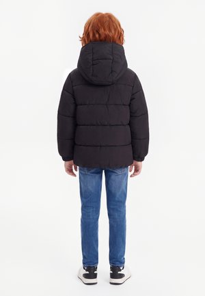 Barn med rødt hår iført en sort hættetrøje med dunjakke, blå jeans og hvide sneakers, der står med ryggen mod på hvid baggrund.