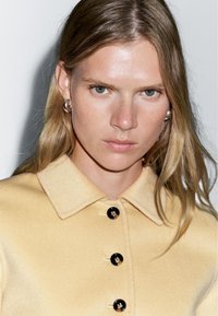 Yellow wool coat with a pointed collar, featuring black buttons. Smooth texture and fitted design with a seamless look.