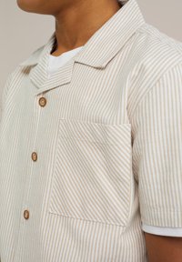 Camicia a maniche corte con bottoni in bianco e strisce beige chiaro, con bottoni in legno e una tasca applicata sul lato sinistro del petto.