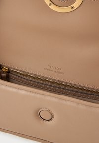 Bolso de mano de cuero beige con un acento circular dorado, bolsillo con cierre de cremallera y nombre de la marca en relieve. Textura suave con un detalle de cierre a presión.