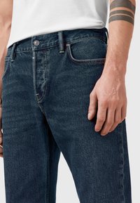 Vaqueros de mezclilla azul oscuro con un diseño ajustado, estilo de cinco bolsillos, cierre de botón de metal y detalles de costura sutiles. Textura suave y ligera elasticidad.