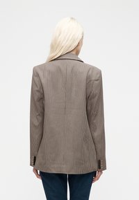 Γυναίκα με μακριά ξανθά μαλλιά, που φοράει καφέ καρό σακάκι και σκούρο μπλε τζιν, στέκεται με την πλάτη προς το μέρος μας μπροστά σε λευκό φόντο.