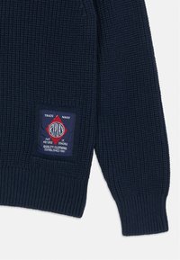 pull en maille bleu marine avec texture côtelée, comportant un patch logo rectangulaire rouge et bleu sur le côté gauche.