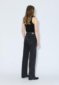 Giovane donna con capelli lunghi, che indossa un top senza maniche nero e jeans neri a vita alta e larghi, in piedi contro un muro bianco.