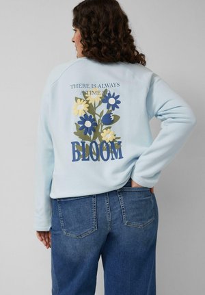 Vrouw met krullend haar, gekleed in een lichtblauwe trui met blauwe en gele bloemen en de tekst "Er is altijd een tijd om te bloeien," gecombineerd met een spijkerbroek.