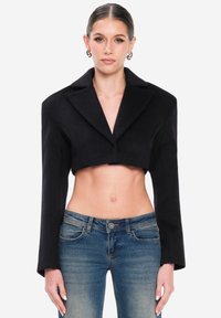 Blazer negro recortado con mangas largas y solapas anchas, hecho de tela suave. Combinado con jeans ajustados azules, mostrando un estilo de abdomen descubierto.