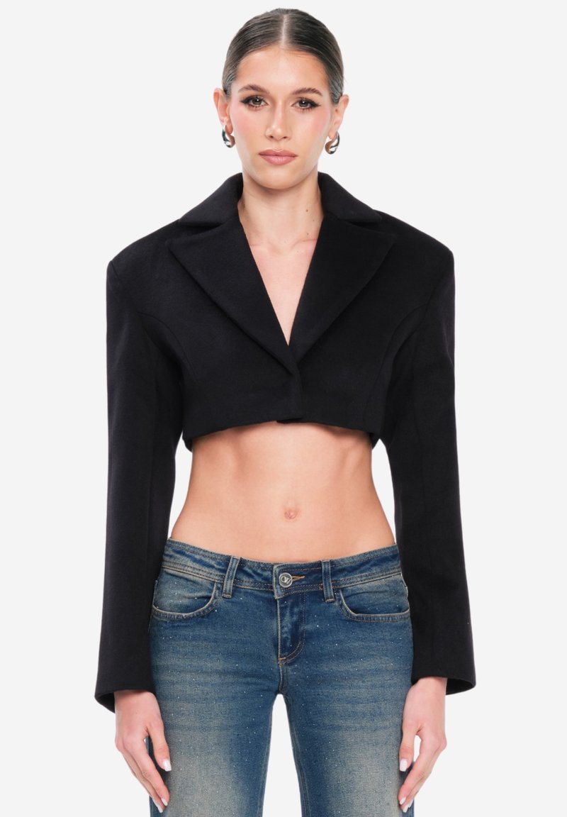 Blazer negro recortado con mangas largas y solapas anchas, hecho de tela suave. Combinado con jeans ajustados azules, mostrando un estilo de abdomen descubierto.