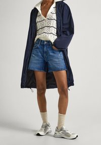 Osoba oblečená v námořnickém kabátu, bílém pleteném svetru s černými pruhy, denim šortkách, béžových ponožkách a bílých teniskách, stojící na světlejším pozadí.