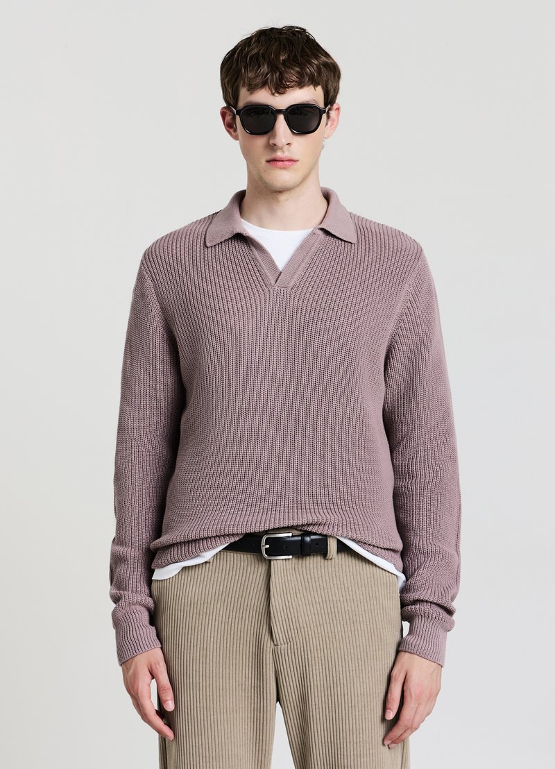 Maglione rosa chiaro lavorato a maglia con collo, caratterizzato da un design a scollo a V. Indossato sopra una camicia bianca e abbinato a pantaloni beige a coste. Cintura nera.