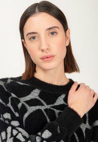 Giovani donna con capelli castani lisci che tocca la spalla, indossando un maglione fuzzy a fantasia nera e grigia, sfondo neutro.