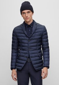 Blazer imbottito blu navy con motivo a righe orizzontali, due bottoni e rever a punta. Indossato sopra una camicia scura, abbinato a un berretto di maglia.
