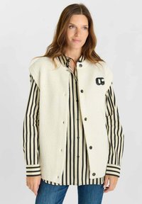Gilet senza maniche in misto lana color crema caratterizzato da una superficie testurizzata, grandi bottoni neri e un logo ricamato. Sotto, una camicia a righe nera e crema.