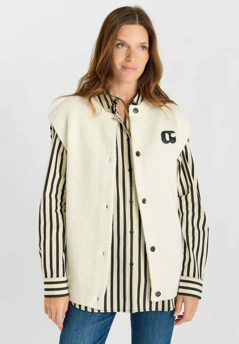 Gilet senza maniche in misto lana color crema caratterizzato da una superficie testurizzata, grandi bottoni neri e un logo ricamato. Sotto, una camicia a righe nera e crema.