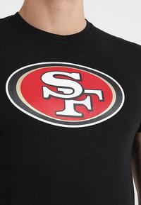 Camiseta de algodón negra con un destacado logo de los San Francisco 49ers en rojo y blanco dentro de una forma ovalada, contorneada en dorado y blanco.