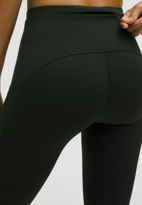 Sötétzöld, sima textúrájú, magas derekú leggings, diszkrét cipzáras zsebbel a hátsó részen és ívelt varrásokkal.