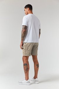 Weißes Baumwoll-T-Shirt, beige Cargo-Shorts mit Seitentaschen und weiße Sneakers. Tattoos an den Beinen sichtbar. Schlichte, funktionale Kleidung.