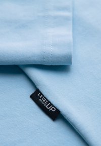 Tissu bleu clair plié avec une étiquette noire visible portant la mention "LEVEL UP" cousue dans la couture.