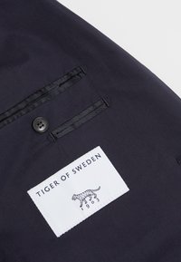 Svart tygjacka med en vänster bröstficka, som har en vit lapp där det står "TIGER OF SWEDEN" och en illustration av en tiger.