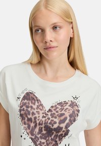 Witte t-shirt met korte mouwen, voorzien van een hartontwerp in luipaardprint, omlijnd met kleine zwarte stippen en speelse krabbels. Zachte stoftextuur.