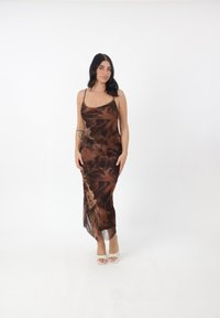 Robe midi brune et transparente avec des motifs floraux et des bretelles fines, affichant une silhouette ajustée et une fente sur le côté. Portée avec des sandales de couleur claire.